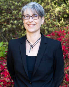 Dr Rebecca Carruthers Den Hoed wearing glasses, black suit jacket and a necklace, standing in front of a colorful leafy background.