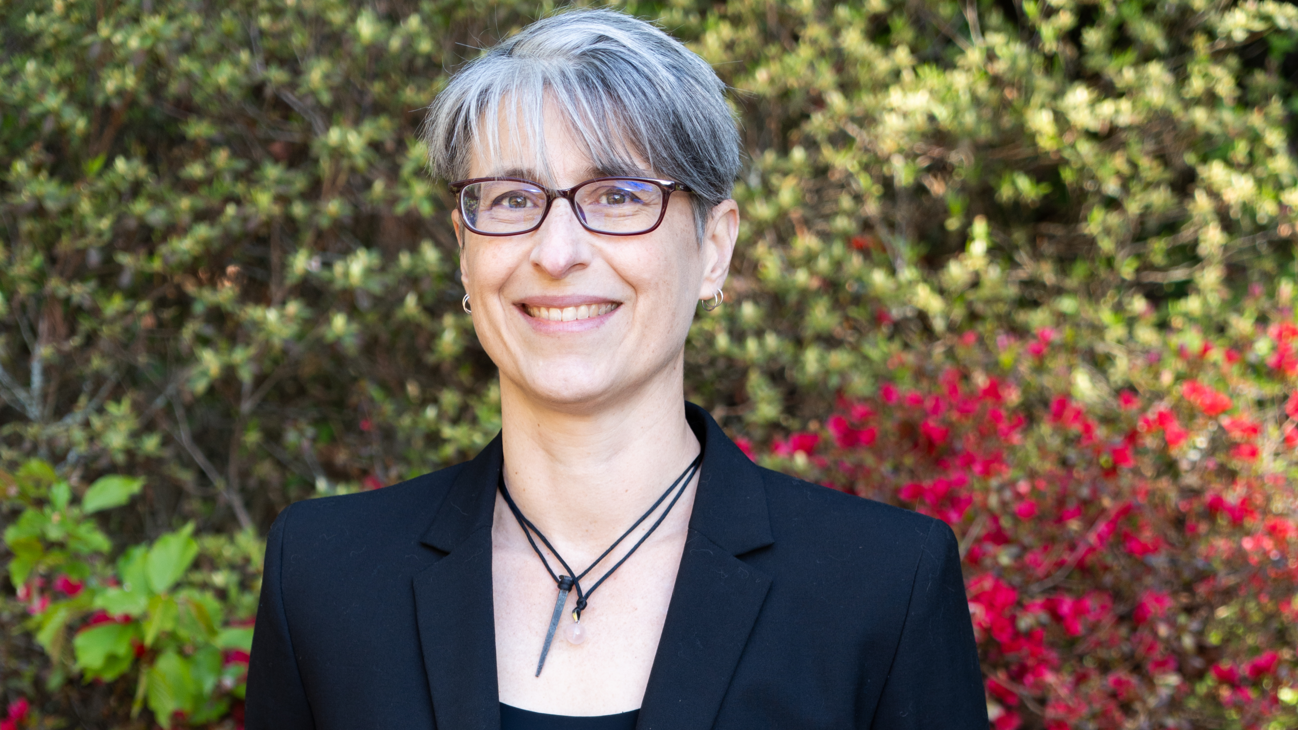 Headshot of Dr Rebecca Carruthers Den Hoed wearing glasses, black suit jacket and a necklace, standing in front of a colorful leafy background.