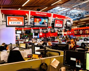 Inside view of a busy newsroom with multiple monitors, and people at their workstations inside their workplace cubicles