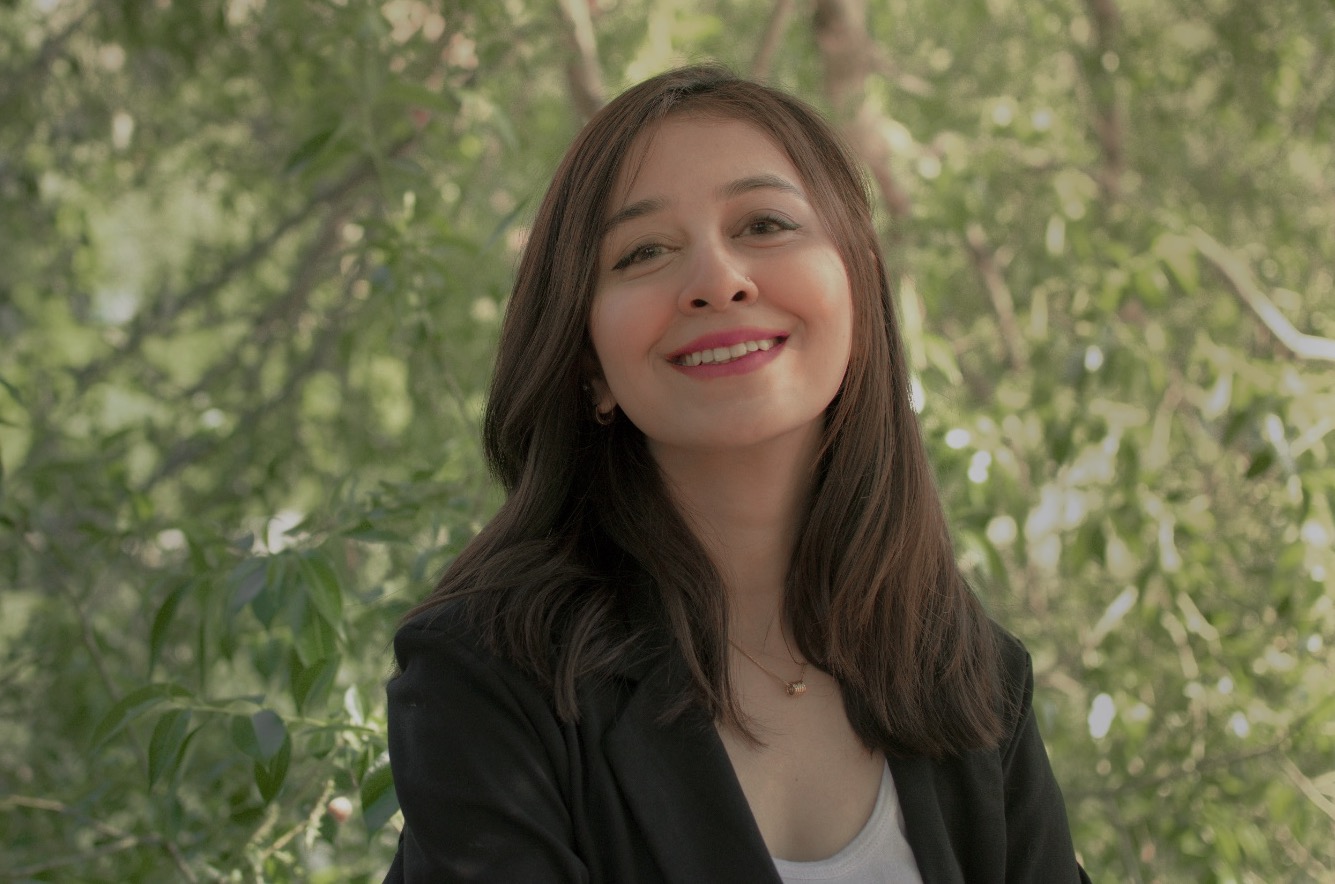 A smiling woman wearing a dark jacket in front of a tree with green leaves