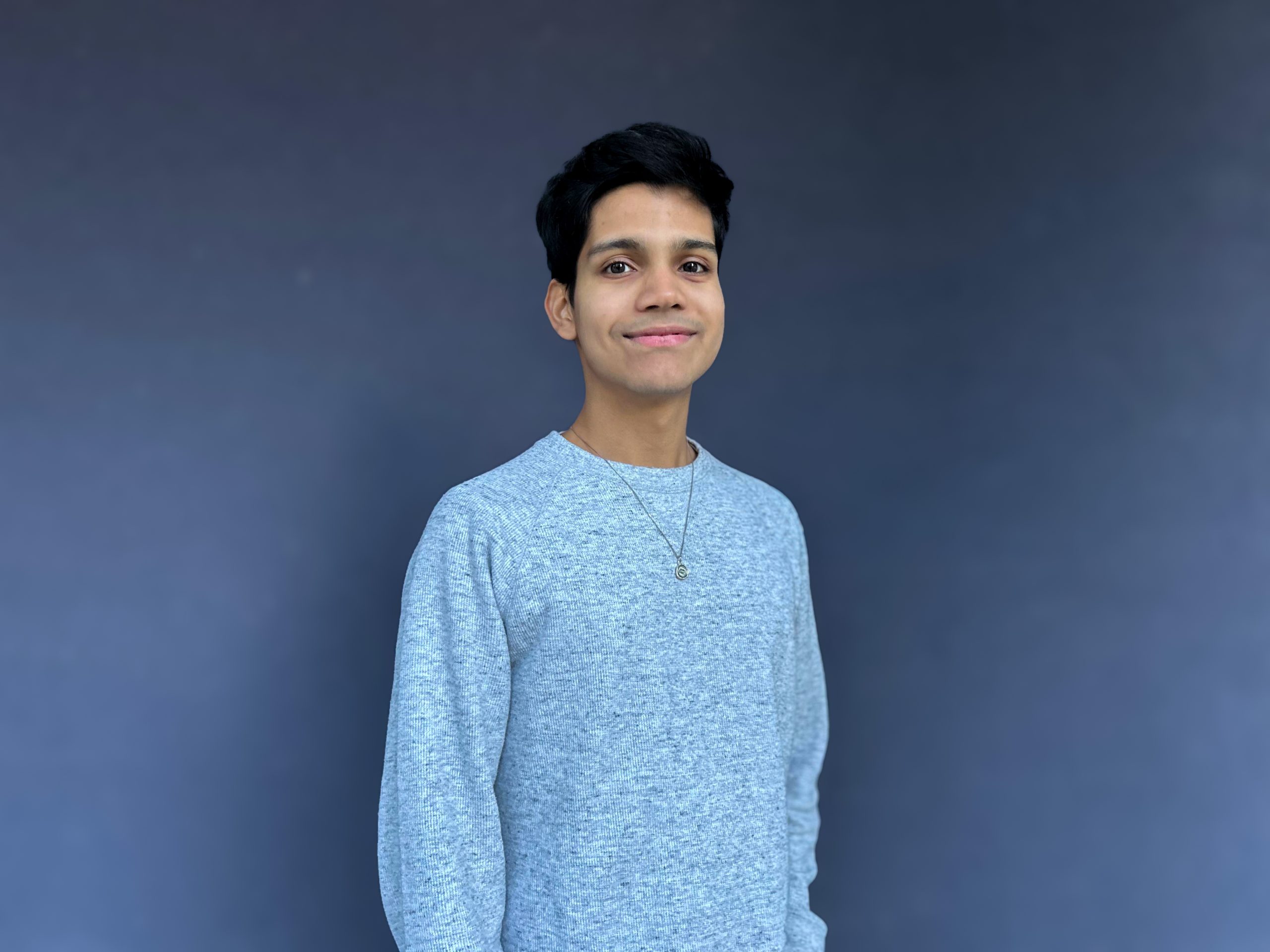 A smiling guy wearing a grey sweatshirt standing against a dark blue background