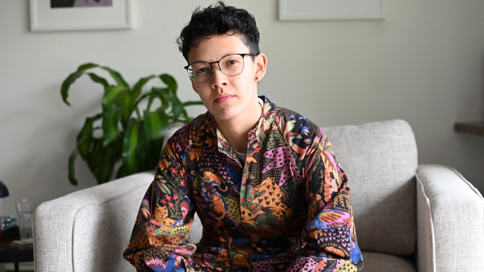 A bespectacled woman wearing a colorful shirt sitting on a beige couch against a wall